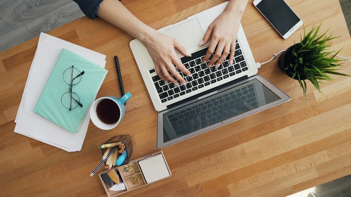Overhead view of hands typing on a laptop at a wooden desk with papers, glasses and coffee