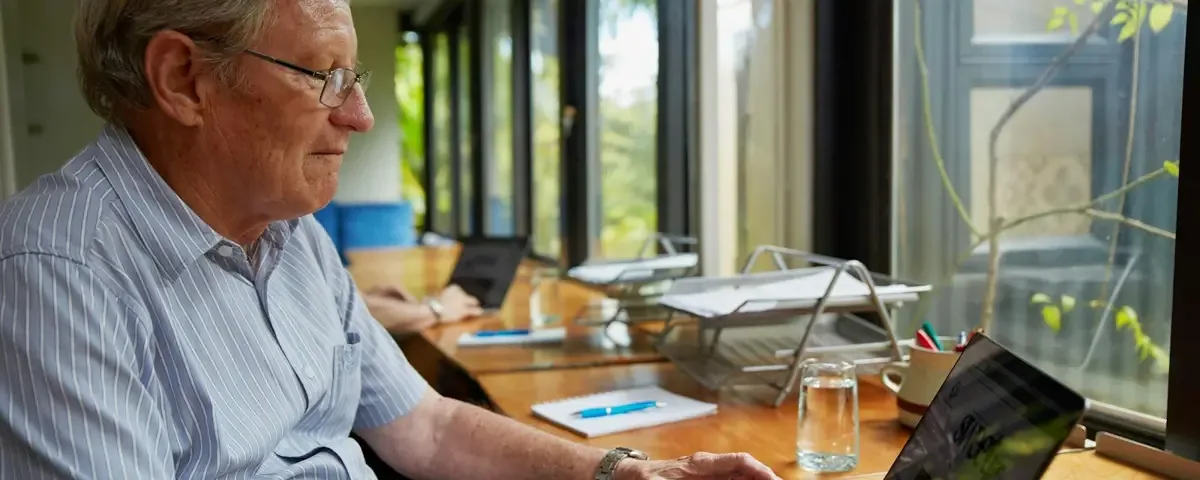 An older man in glasses types on a laptop at a desk beside a bright window
